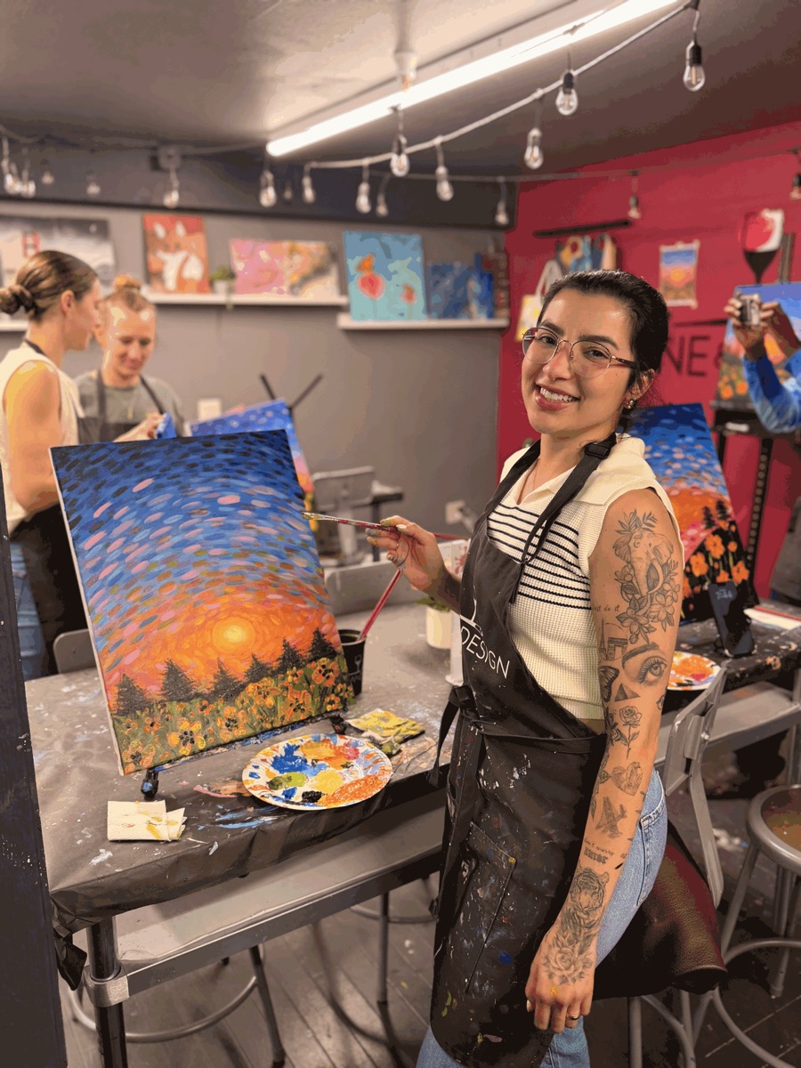 Maysa Tavares smiling beside a sunset painting at her easel